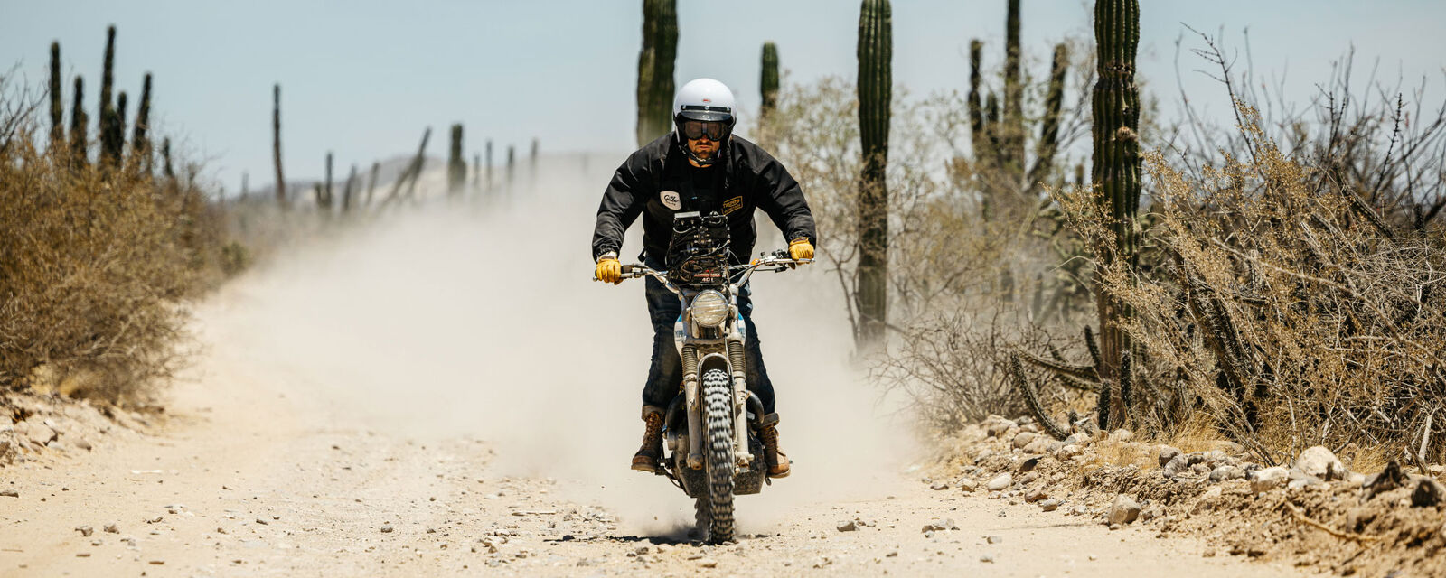 A rider in a white Bell helmet kicking up dust on a desert dirt road lined with cactus.