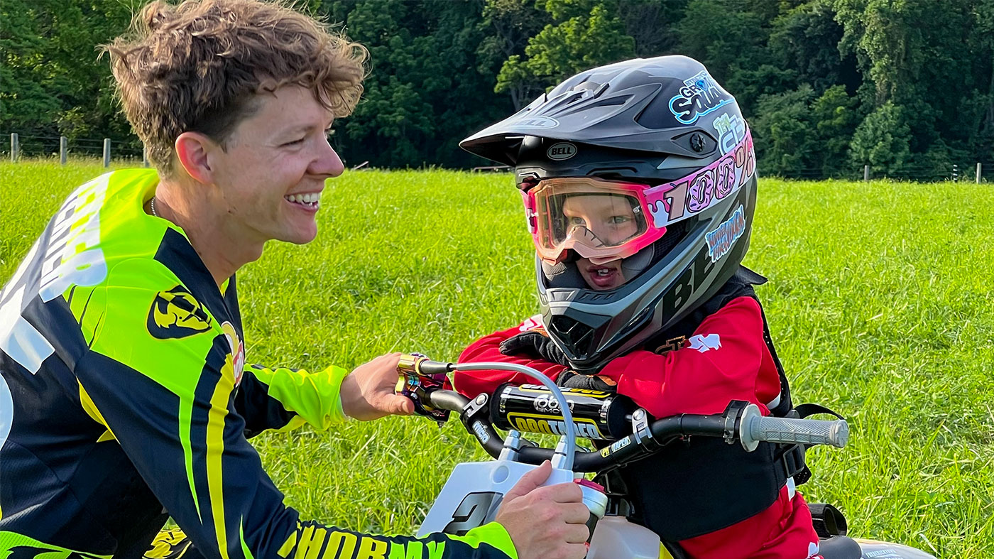 Cory smiling at his son who is sitting on a motorcyle with his helmet and gear on.