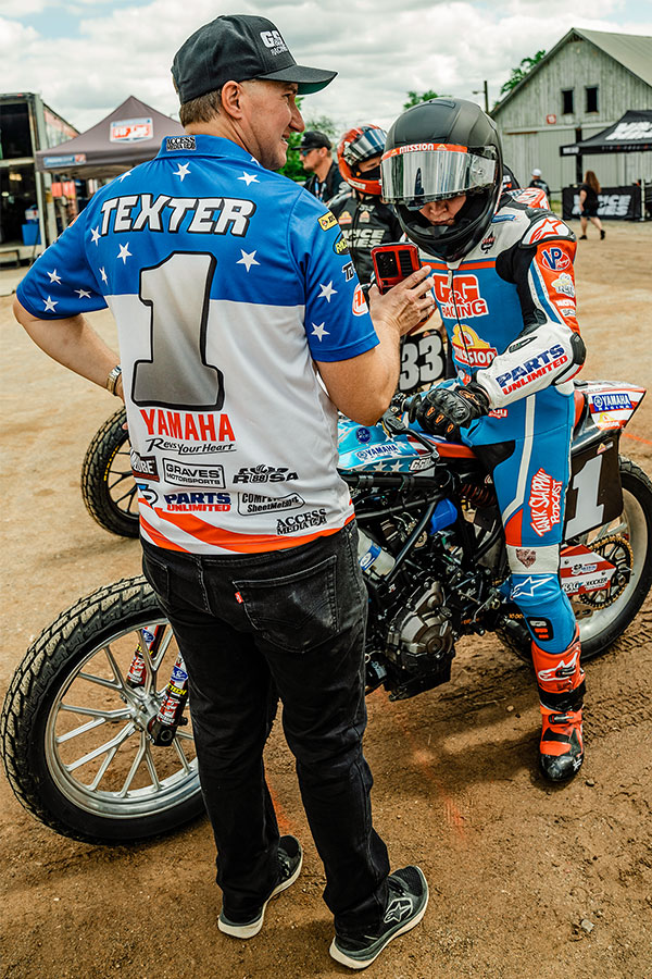 A team member holding up a phone for Cory to read while sitting on his motorcycle ready to race.